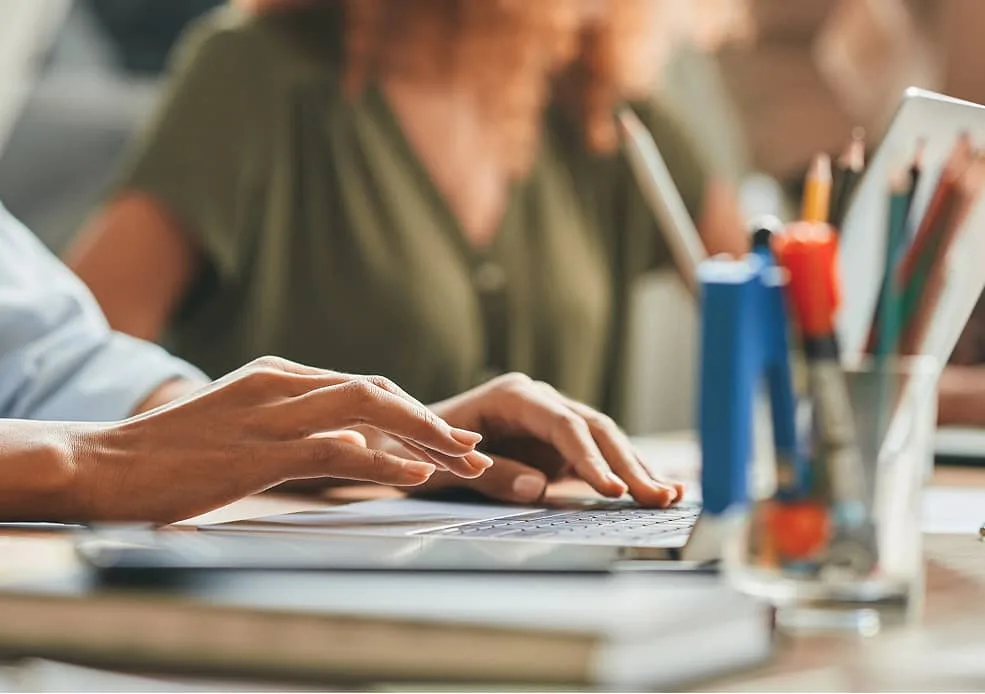 Person tippt auf Laptop, weitere Person arbeitet im Hintergrund