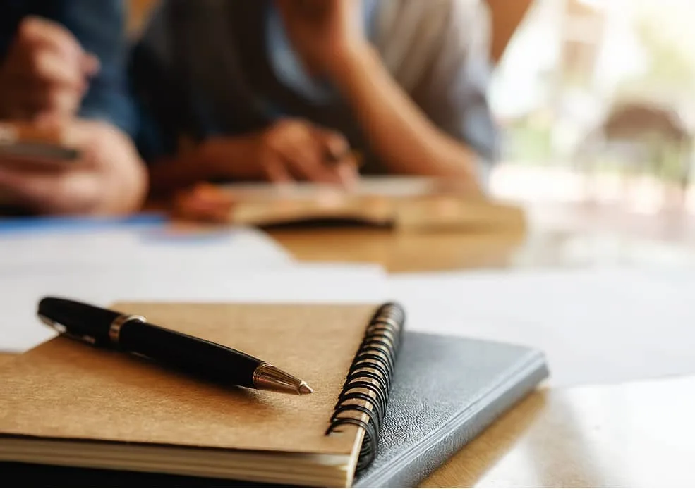 Notizbuch mit Stift auf Tisch, im Hintergrund arbeitende Personen
