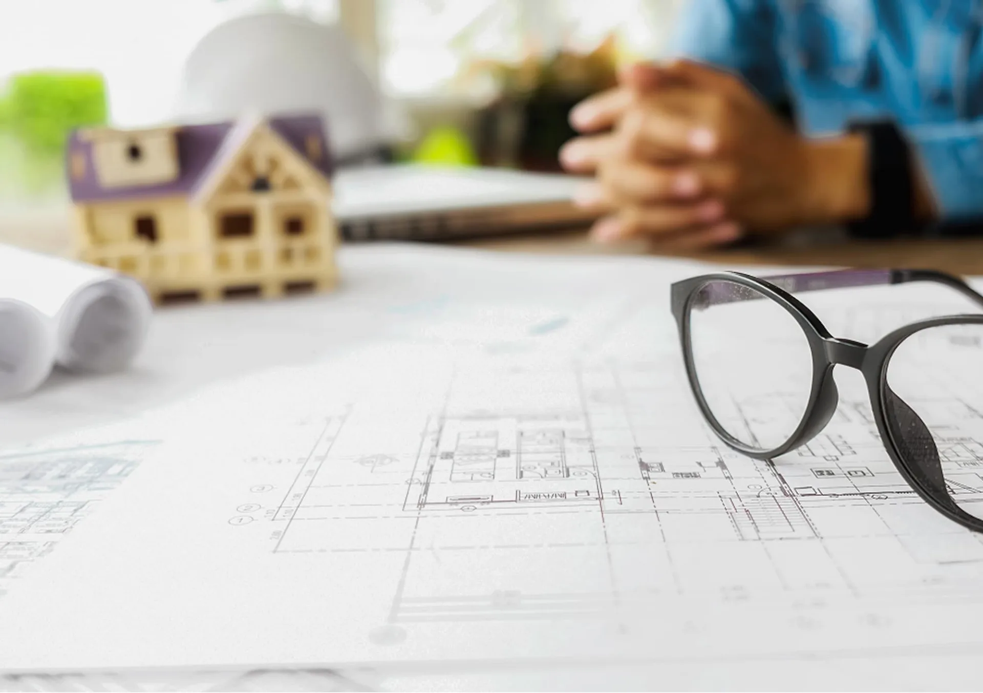 `Architekturpläne auf Tisch mit Brille, Hausmodell und Person im Hintergrund`